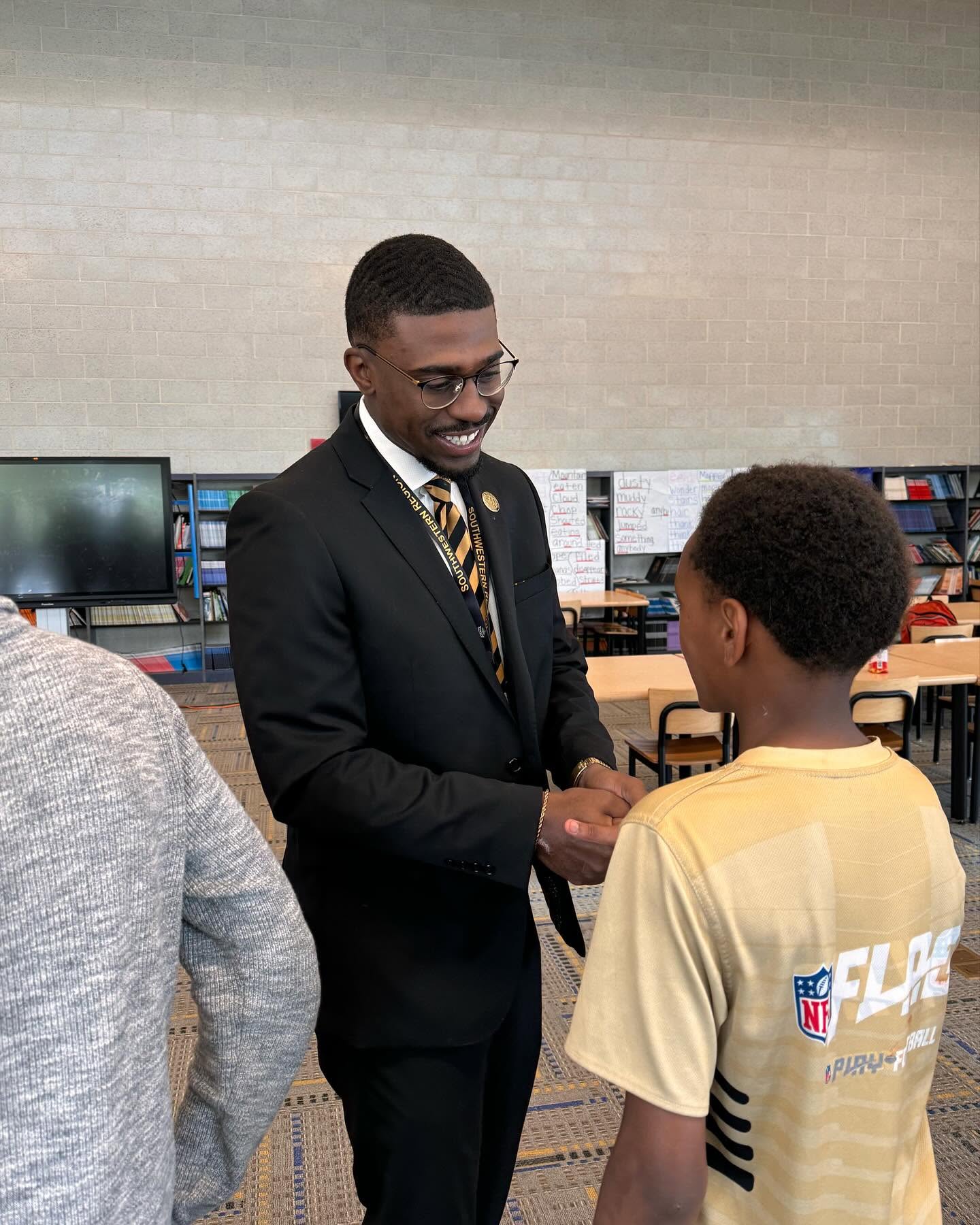Xi Tau Lambda brother in a suit greeting a student with a handshake during a school mentorship visit