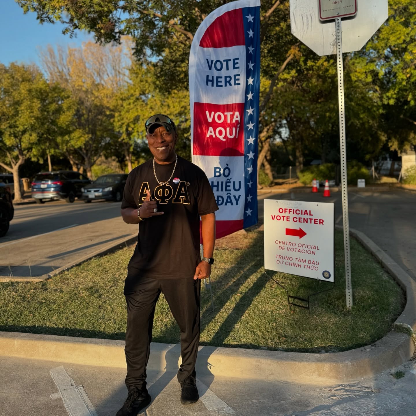 Xi Tau Lambda brother in Alpha Phi Alpha gear at an official vote center with a Vote Here banner
